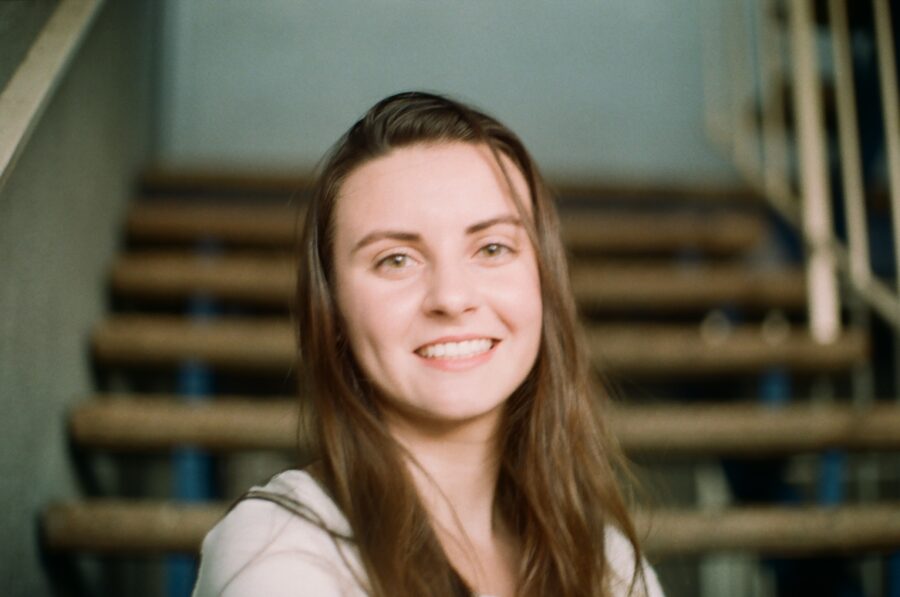 A close-up of Alex's face, her long brown hair draping past her neck beyond the lower frame of the photo. She's slightly blurred, but has a vibrant and youthful smile. Behind her, an out-of-focus, modern architecture wooden staircase leads up into a concrete area, with a white metal railing to the side.